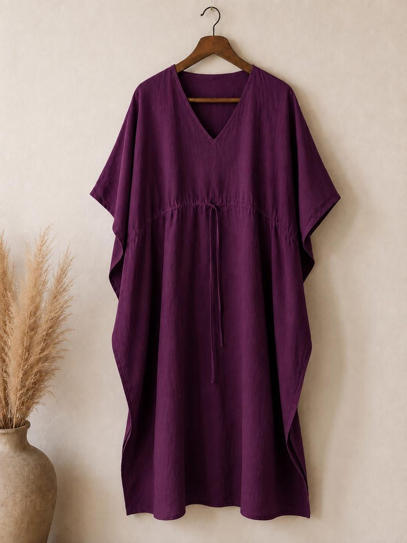 Puede incluir: Un vestido caft&aacute;n de color morado intenso con escote en V, mangas cortas anchas y cintura con cord&oacute;n ajustable y finas cintas a juego. La prenda se muestra en una percha de madera, exhibiendo una silueta holgada y fluida de tela mate.