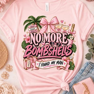 May include: A light pink t-shirt featuring a graphic print with the text "NO MORE BOMBSHELLS" and "I FOUND MY MAN". The design includes a palm tree, a pink gingham bow, champagne glasses, a bottle, heart-shaped sunglasses, and a suitcase.