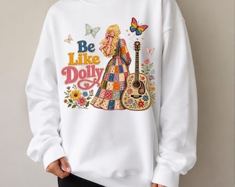May include: A white long-sleeve sweatshirt featuring a graphic print of a person with blonde hair in a patchwork dress standing next to an acoustic guitar. The text "Be Like Dolly" is printed in blue and pink, surrounded by butterflies and flowers.