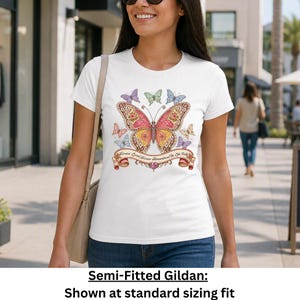 Op de afbeelding: Een wit T-shirt met korte mouwen en een grote, gedetailleerde vlinderprint in roze, gele en oranje tinten, omringd door kleinere vlinders. Een decoratieve banner onder de centrale vlinder bevat de tekst Forever Loved, Forever Remembered, Be Like Dolly.
