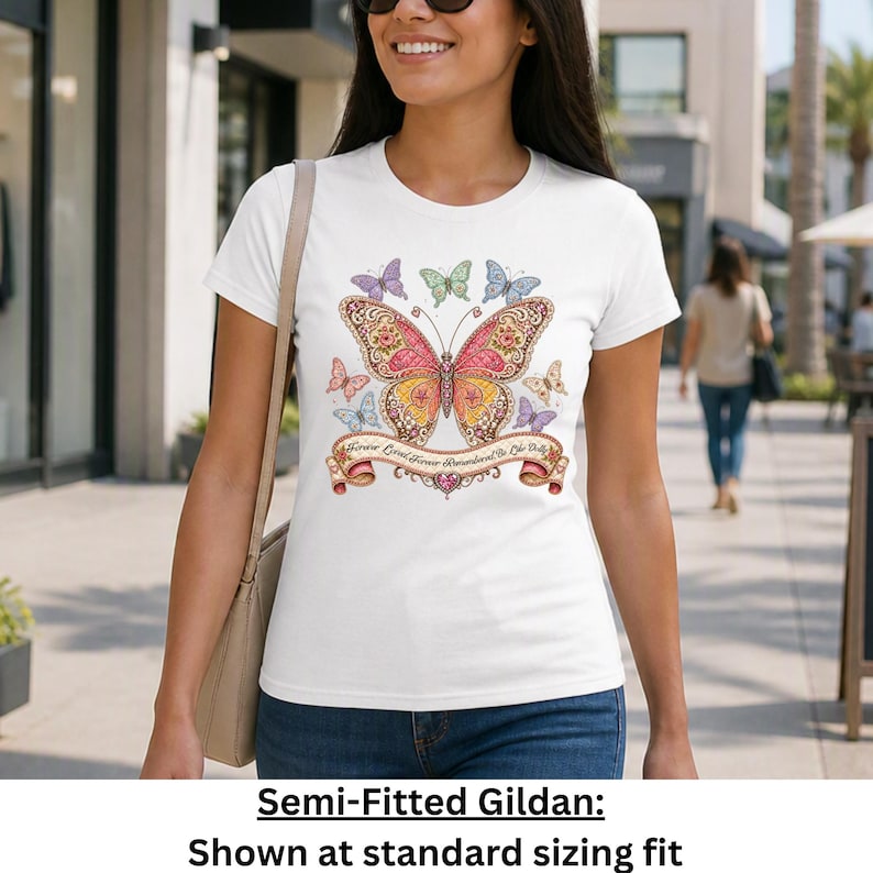 Op de afbeelding: Een wit T-shirt met korte mouwen en een grote, gedetailleerde vlinderprint in roze, gele en oranje tinten, omringd door kleinere vlinders. Een decoratieve banner onder de centrale vlinder bevat de tekst Forever Loved, Forever Remembered, Be Like Dolly.