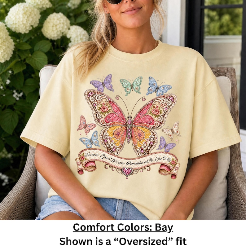 Op de afbeelding: Een lichtgeel t-shirt met korte mouwen en een grote vlinderprint in het midden met roze, oranje en gouden vleugels. Kleinere vlinders omringen het ontwerp. Een banner eronder bevat de tekst Forever Loved, Forever Remembered, Be Like Dolly.