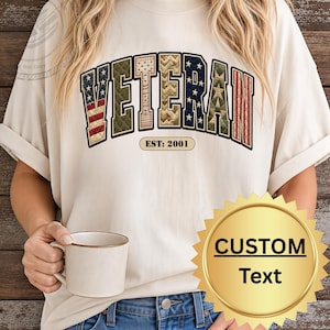 May include: A cream-colored t-shirt featuring the word VETERAN in large, block-style letters with various patterns including American flag motifs, stars, and quilted textures. Below the word, the text EST: 2001 is printed in a small, rounded rectangle.