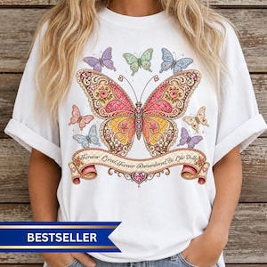 May include: A white t-shirt featuring a large, central butterfly graphic with pink, orange, and yellow wings adorned with floral patterns. Smaller butterflies surround the center design. A banner below reads, Forever Loved, Forever Remembered, Be Like Dolly.