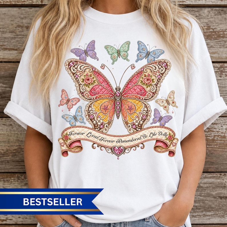 Op de afbeelding: Een wit t-shirt met een grote, centrale vlinderprint met roze, oranje en gele vleugels versierd met bloemmotieven. Kleinere vlinders omringen het ontwerp. Een banner eronder bevat de tekst, Forever Loved, Forever Remembered, Be Like Dolly.