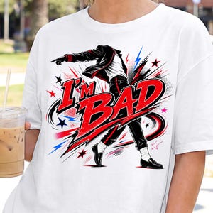 May include: A white t-shirt featuring a graphic illustration of a dancer in a black jacket and trousers with white socks and black shoes. The text I'M BAD is printed in large red letters across the center, surrounded by red and blue star accents.