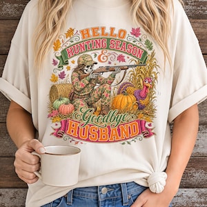 May include: A cream-colored t-shirt featuring a graphic of a skeleton in camouflage clothing holding a rifle, surrounded by autumn leaves, pumpkins, a hay bale, and a turkey. The text reads "HELLO HUNTING SEASON & goodbye HUSBAND".