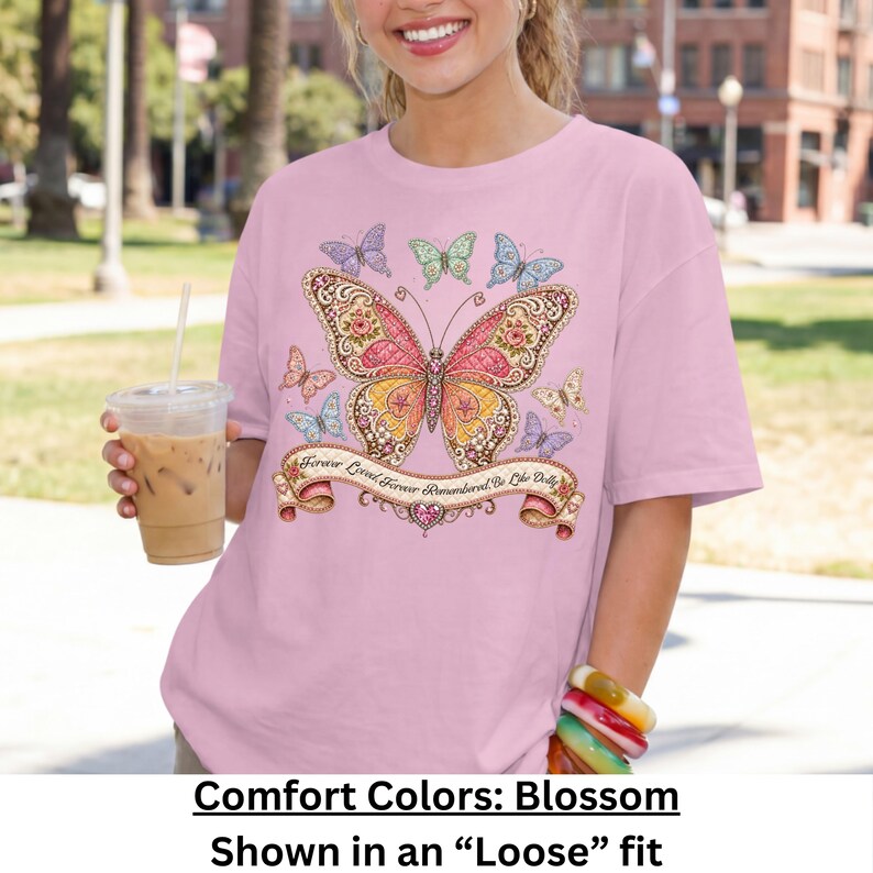 Op de afbeelding: Een lichtroze t-shirt met een grote, gedetailleerde vlinderprint in roze, oranje en goudtinten. Het ontwerp bevat kleinere vlinders en een decoratieve banner met de tekst, Forever Loved, Forever Remembered, Be Like Dolly.