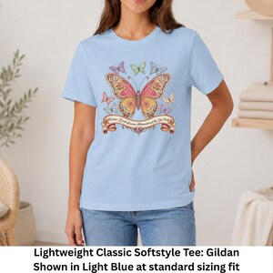Op de afbeelding: Een lichtblauw t-shirt met korte mouwen en een grote centrale vlindergrafiek met ingewikkelde roze, oranje en gele patronen. Kleinere vlinders omringen het midden en een banner eronder bevat de tekst Forever Loved Forever Remembered Be Like Dolly.