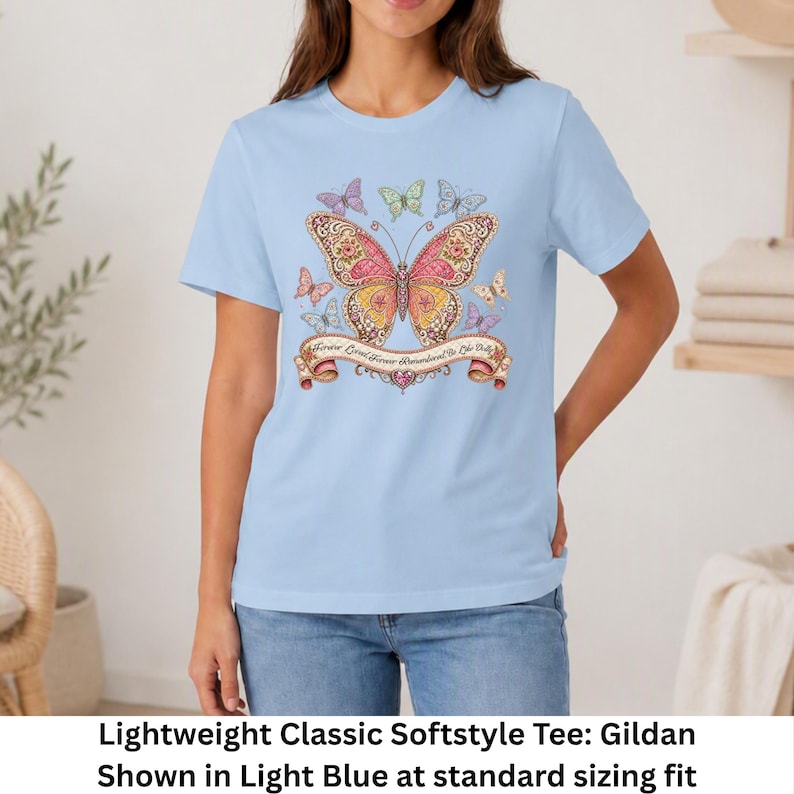 Op de afbeelding: Een lichtblauw t-shirt met korte mouwen en een grote centrale vlindergrafiek met ingewikkelde roze, oranje en gele patronen. Kleinere vlinders omringen het midden en een banner eronder bevat de tekst Forever Loved Forever Remembered Be Like Dolly.