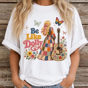 May include: A white t-shirt featuring a graphic print of a person with blonde hair wearing a patchwork dress, standing next to an acoustic guitar. The design includes colorful flowers, butterflies, and the text Be Like Dolly in blue and pink.