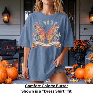 Op de afbeelding: Een stoffig blauw T-shirt met korte mouwen en een grote centrale vlindergrafiek met ingewikkelde roze, gele en oranje patronen. Kleinere vlinders omringen het midden en een banner eronder bevat de tekst Forever Loved Forever Remembered Be Like Dolly.