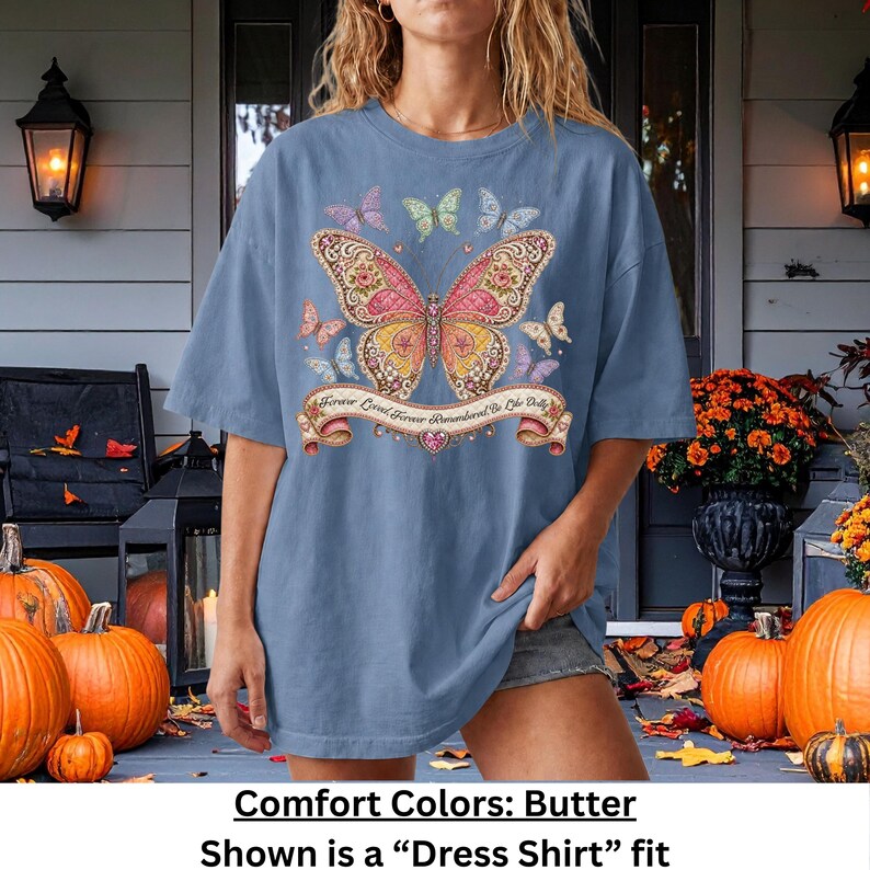 Op de afbeelding: Een stoffig blauw T-shirt met korte mouwen en een grote centrale vlindergrafiek met ingewikkelde roze, gele en oranje patronen. Kleinere vlinders omringen het midden en een banner eronder bevat de tekst Forever Loved Forever Remembered Be Like Dolly.