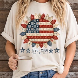 May include: A cream-colored t-shirt featuring a large sunflower graphic with petals and a center designed like a patchwork American flag. Below the sunflower, the text 1776-2026 is printed, flanked by two small stars on each side and a tiny star below.