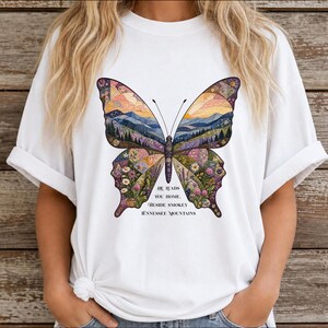 May include: A white short-sleeved t-shirt featuring a large butterfly graphic. The butterfly wings contain a landscape of mountains, forests, and flowers in a quilt-like pattern. Below the butterfly, the text reads: HE LEADS YOU HOME BESIDE SMOKEY TENNESSEE MOUNTAINS.