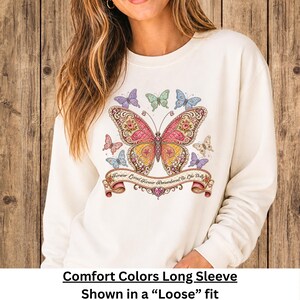 Op de afbeelding: Een cr&egrave;mekleurig sweatshirt met lange mouwen en een grote, gedetailleerde vlinderprint in roze, gele en gouden tinten. Onder de vlinder bevindt zich een decoratieve banner met de tekst "Forever Loved, Forever Remembered, Be Like Dolly."