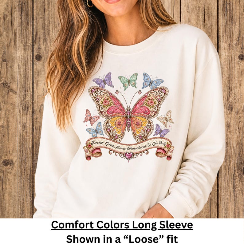 Op de afbeelding: Een cr&egrave;mekleurig sweatshirt met lange mouwen en een grote, gedetailleerde vlinderprint in roze, gele en gouden tinten. Onder de vlinder bevindt zich een decoratieve banner met de tekst "Forever Loved, Forever Remembered, Be Like Dolly."
