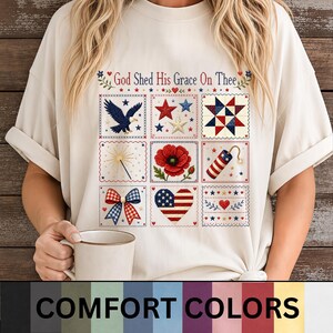 May include: A cream-coloured t-shirt featuring a grid of nine embroidered-style patriotic illustrations, including an eagle, stars, a quilt block, a sparkler, a poppy, a firecracker, a bow, a heart flag, and the text God Shed His Grace On Thee.