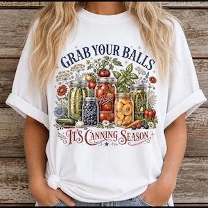 May include: A white t-shirt featuring a graphic print of four glass jars filled with various preserved vegetables and fruits, surrounded by botanical illustrations. The text reads GRAB YOUR BALLS and IT'S CANNING SEASON in dark blue and red.