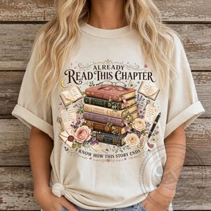May include: A cream-colored t-shirt featuring a graphic of a stack of five books with reading glasses on top. The text reads ALREADY READ THIS CHAPTER and I KNOW HOW THIS STORY ENDS. The design includes floral accents, a fountain pen, and letters.