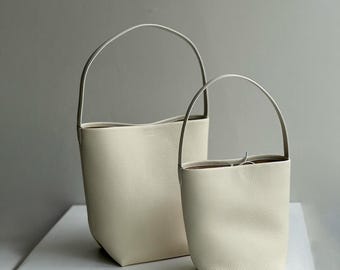 May include: Two off-white leather bucket bags of different sizes. Each bag has a single, arched handle and a textured surface. The bags are set against a neutral background, highlighting their minimalist design and neutral color.
