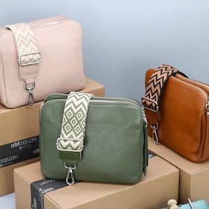 Könnte beinhalten: Drei Leder-Umhängetaschen in Rosa, Olivgrün und Braun, jede mit einem gemusterten Riemen und silberfarbenen Beschlägen. Die Taschen werden auf Pappkartons präsentiert.