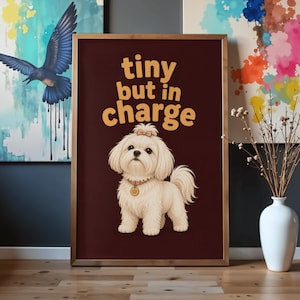 May include: A framed wall art print with a dark brown background featuring a fluffy cream-colored dog with a small bow on its head and a gold tag on its collar. The text "tiny but in charge" is printed in large, bold, light orange letters above the dog.