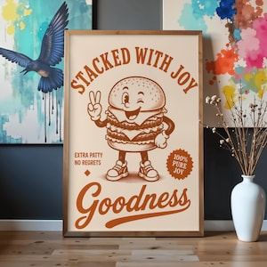 May include: A framed print featuring a cartoon burger character winking and giving a peace sign. The text reads "Stacked with Joy," "Extra Patty No Regrets," and "Goodness." The burger is brown and white, set against a cream background.