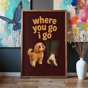 Puede incluir: Un cuadro rectangular con marco sobre un fondo granate oscuro que presenta el texto "where you go i go" en letras amarillas. Debajo del texto hay una ilustración de un perro pequeño, esponjoso y de color marrón claro mirando hacia unas piernas.