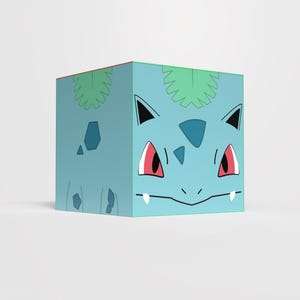 Puede incluir: Un cubo azul claro con la cara de Bulbasaur, un personaje popular de la franquicia Pokémon. El cubo tiene ojos rojos, orejas negras y un diseño de hojas verdes en la parte superior. El diseño es una ilustración de estilo caricatura.