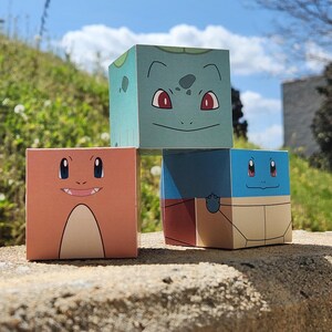 Puede incluir: Tres figuras de papel cuadradas y coloridas de personajes de dibujos animados populares. Las figuras son naranjas, azules y verdes, cada una con rasgos faciales distintos. Están dispuestas sobre una superficie de piedra contra un fondo de vegetación y cielo azul.