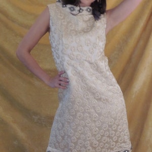 Puede incluir: Un vestido blanco sin mangas con cuello alto y un patr&oacute;n de remolino. El vestido tiene un ribete de cuentas alrededor del cuello y el dobladillo.