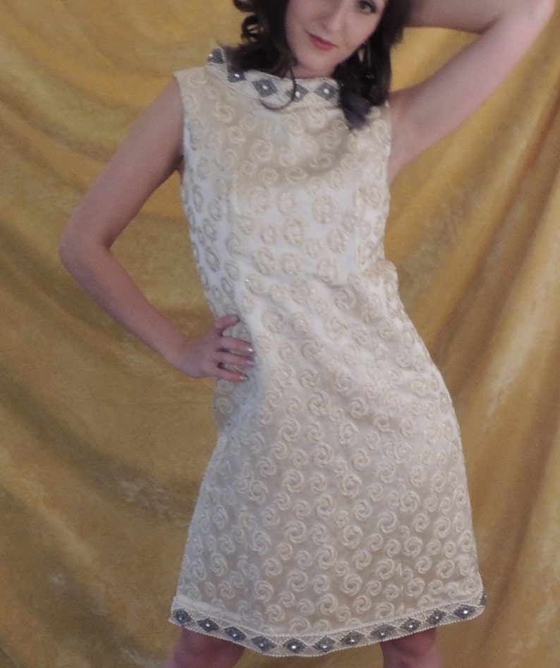 Puede incluir: Un vestido blanco sin mangas con cuello alto y un patr&oacute;n de remolino. El vestido tiene un ribete de cuentas alrededor del cuello y el dobladillo.