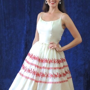 May include: A white dress with red floral embroidery. The dress has a fitted bodice and a full skirt. The model is wearing the dress with a simple hairstyle and earrings.