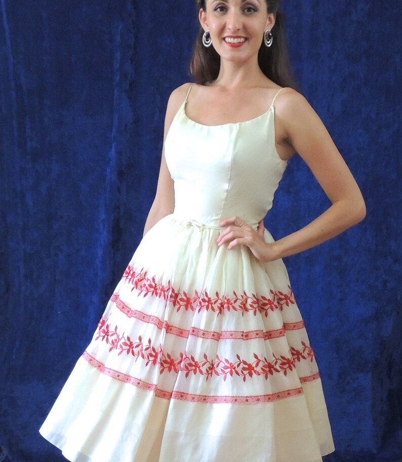 May include: A white dress with red floral embroidery. The dress has a fitted bodice and a full skirt. The model is wearing the dress with a simple hairstyle and earrings.