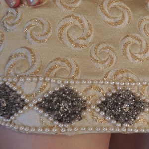 Puede incluir: Una falda de color crema con un patr&oacute;n de espiral dorado y un ribete de cuentas. El ribete est&aacute; hecho de peque&ntilde;as perlas blancas y cuentas plateadas.