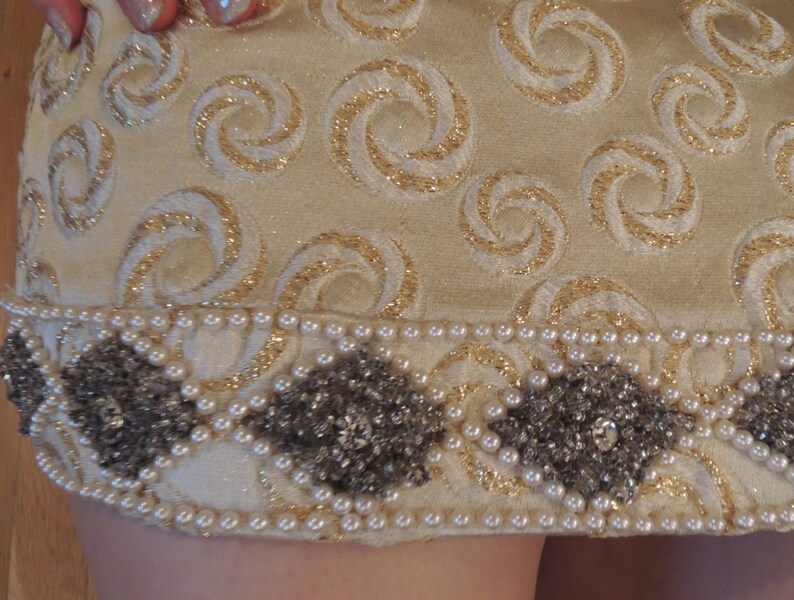 Puede incluir: Una falda de color crema con un patr&oacute;n de espiral dorado y un ribete de cuentas. El ribete est&aacute; hecho de peque&ntilde;as perlas blancas y cuentas plateadas.