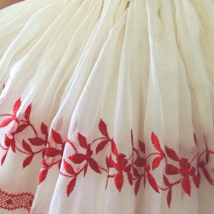 May include: A white dress with red embroidered floral details around the hem. The dress has a gathered waist and a bow at the back.