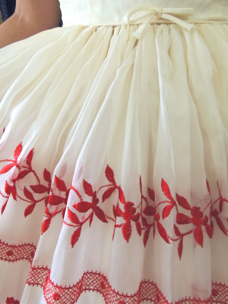 May include: A white dress with red embroidered floral details around the hem. The dress has a gathered waist and a bow at the back.