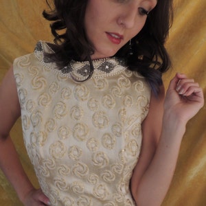 Puede incluir: Un vestido blanco sin mangas con un patr&oacute;n de espiral dorado y un escote con cuentas. El vestido est&aacute; hecho de una tela sedosa y tiene un corpi&ntilde;o ajustado.