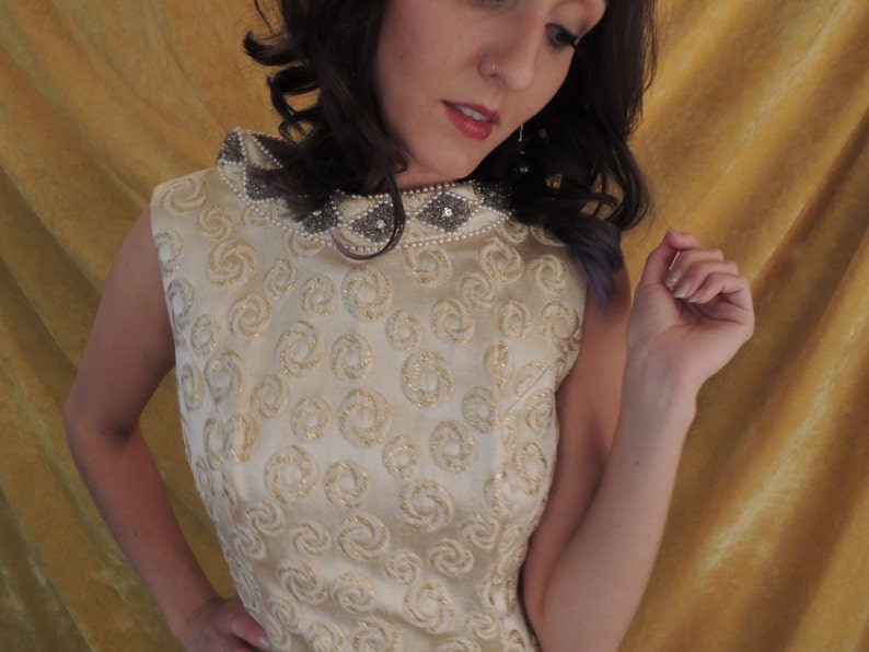 Puede incluir: Un vestido blanco sin mangas con un patr&oacute;n de espiral dorado y un escote con cuentas. El vestido est&aacute; hecho de una tela sedosa y tiene un corpi&ntilde;o ajustado.