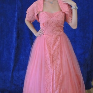 Puede incluir: Vestido rosa coral de estilo vintage con un bolero de encaje a juego. El vestido presenta un escote coraz&oacute;n y una falda de tul completa. El bolero tiene mangas cortas. El vestido es adecuado para un evento formal.