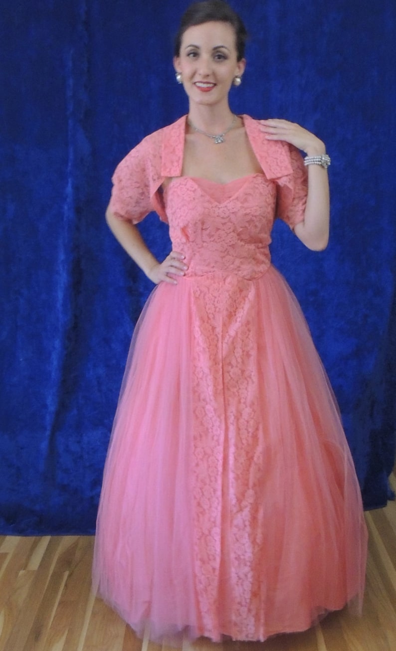 Puede incluir: Vestido rosa coral de estilo vintage con un bolero de encaje a juego. El vestido presenta un escote coraz&oacute;n y una falda de tul completa. El bolero tiene mangas cortas. El vestido es adecuado para un evento formal.