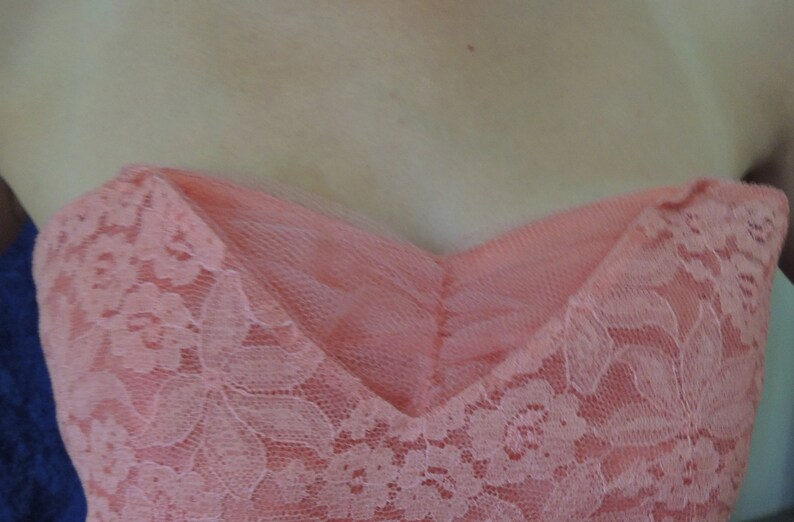 Puede incluir: Un vestido sin tirantes de color coral con escote coraz&oacute;n. El vestido presenta una delicada superposici&oacute;n de encaje floral, que a&ntilde;ade textura y detalles. El corpi&ntilde;o es entallado y el vestido parece estar hecho de una tela suave y fluida.