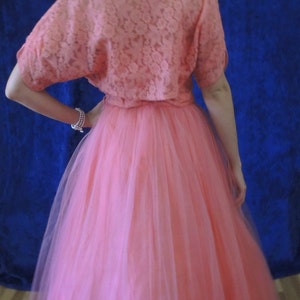 Puede incluir: Vestido rosa coral de estilo vintage. El vestido presenta una parte superior de encaje con mangas cortas y una falda de tul amplia. El conjunto se encuentra sobre un fondo azul oscuro, creando un contraste llamativo.