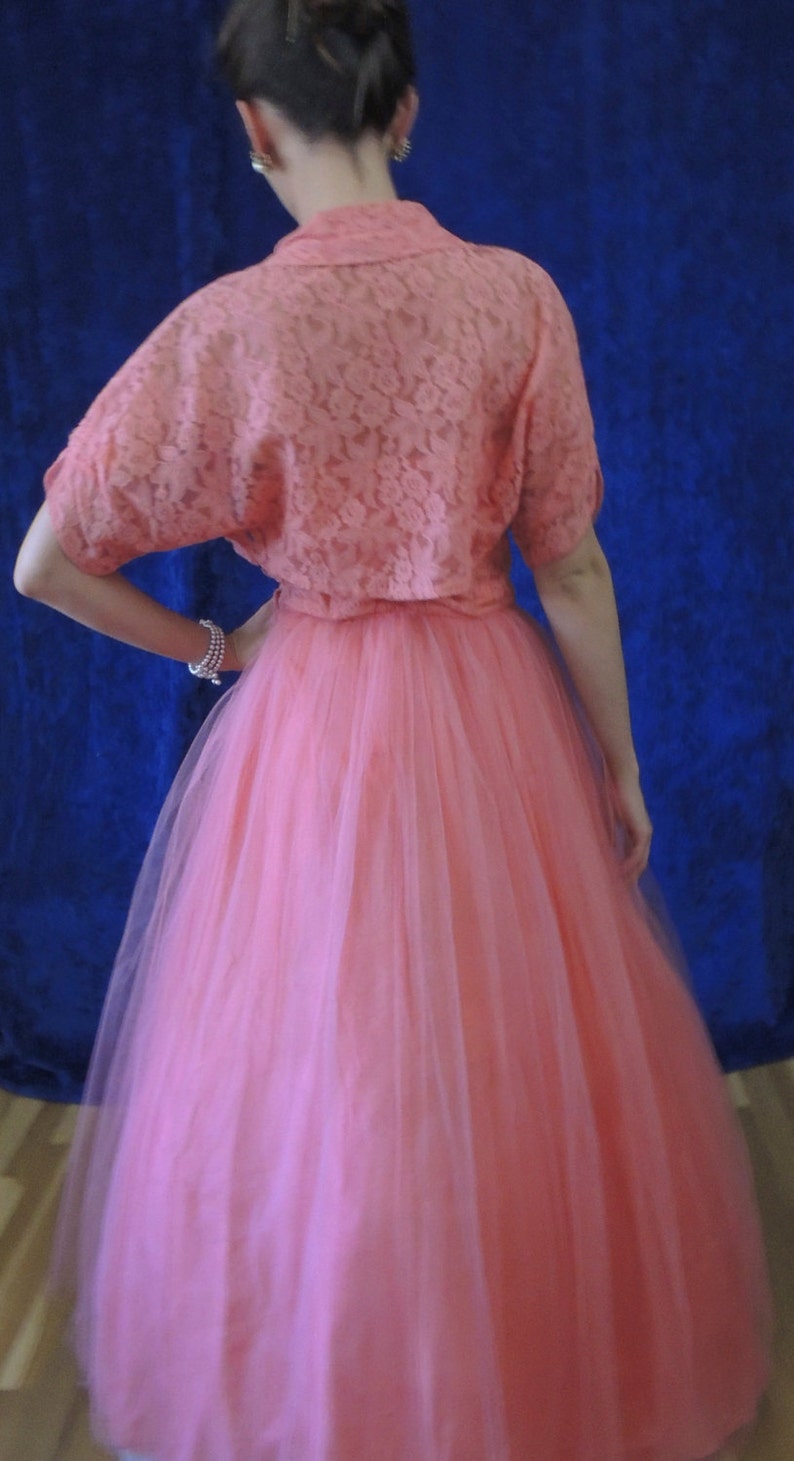 Puede incluir: Vestido rosa coral de estilo vintage. El vestido presenta una parte superior de encaje con mangas cortas y una falda de tul amplia. El conjunto se encuentra sobre un fondo azul oscuro, creando un contraste llamativo.