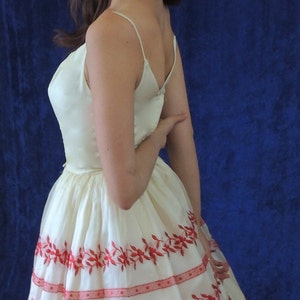 May include: A white dress with red embroidered floral details. The dress has a fitted bodice and a full skirt. The model is wearing silver hoop earrings.
