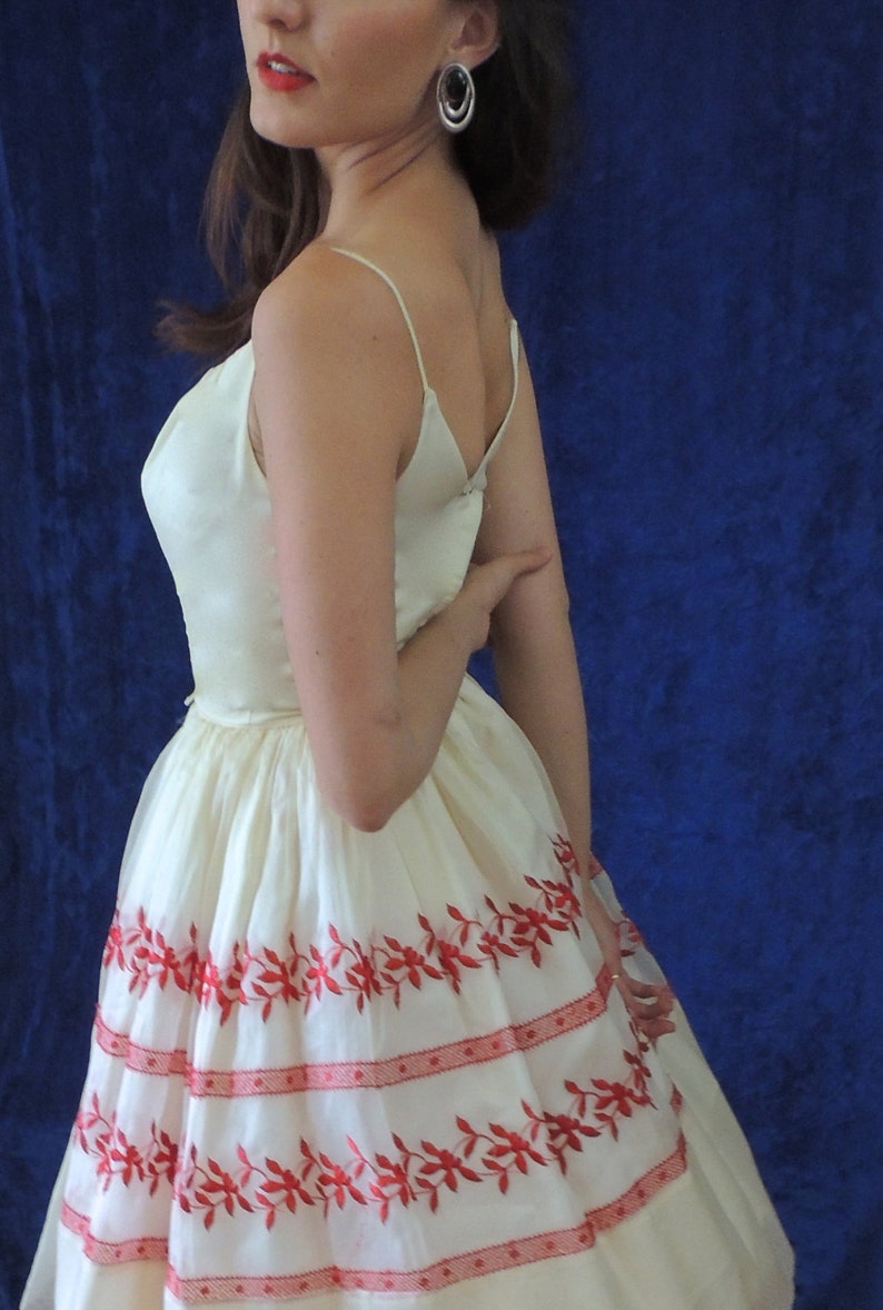 May include: A white dress with red embroidered floral details. The dress has a fitted bodice and a full skirt. The model is wearing silver hoop earrings.