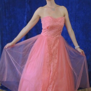 Puede incluir: Un vestido de fiesta color coral sin tirantes. El corpi&ntilde;o presenta una superposici&oacute;n de encaje, y la falda es de tul fluido. El vestido largo crea una silueta elegante. El fondo es azul oscuro, contrastando con el vestido.
