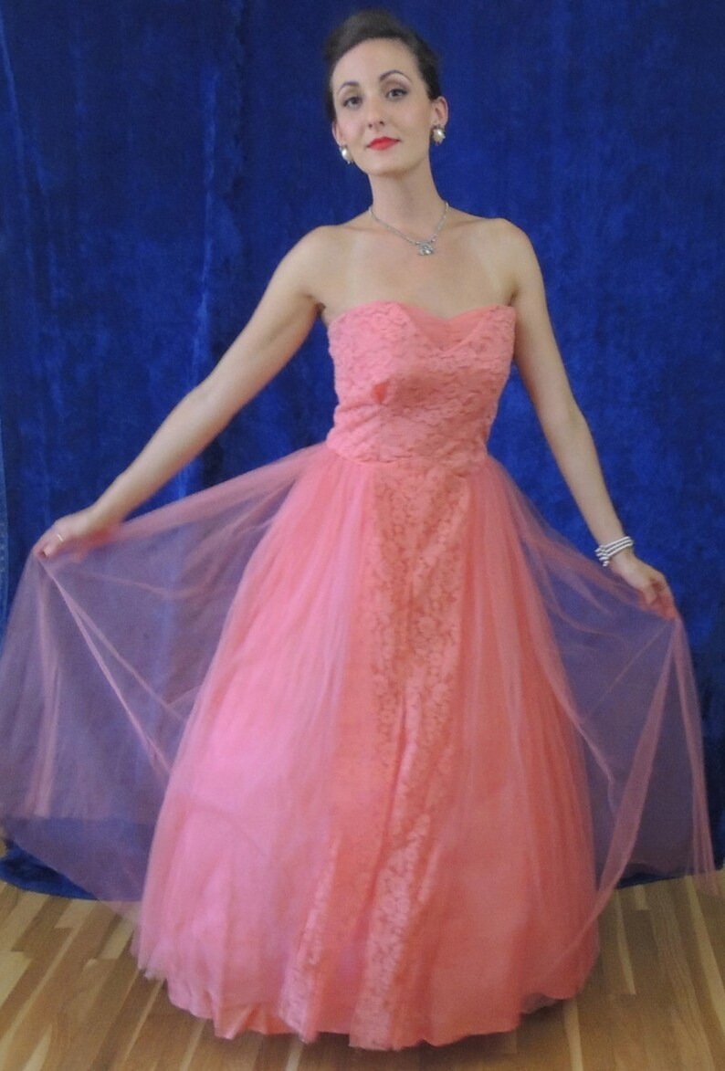 Puede incluir: Un vestido de fiesta color coral sin tirantes. El corpi&ntilde;o presenta una superposici&oacute;n de encaje, y la falda es de tul fluido. El vestido largo crea una silueta elegante. El fondo es azul oscuro, contrastando con el vestido.